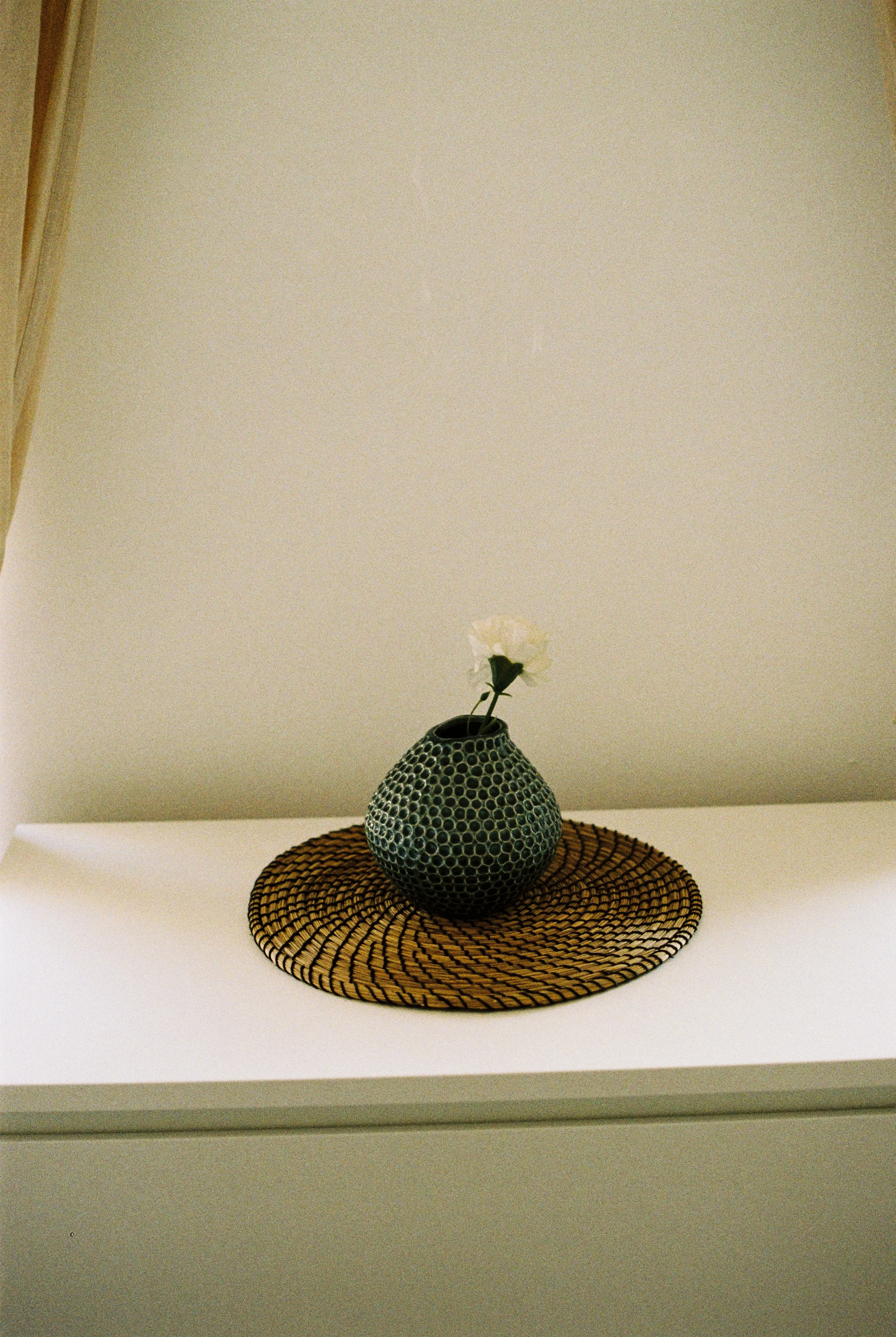 A single carnation in a patterned vase on a woven mat